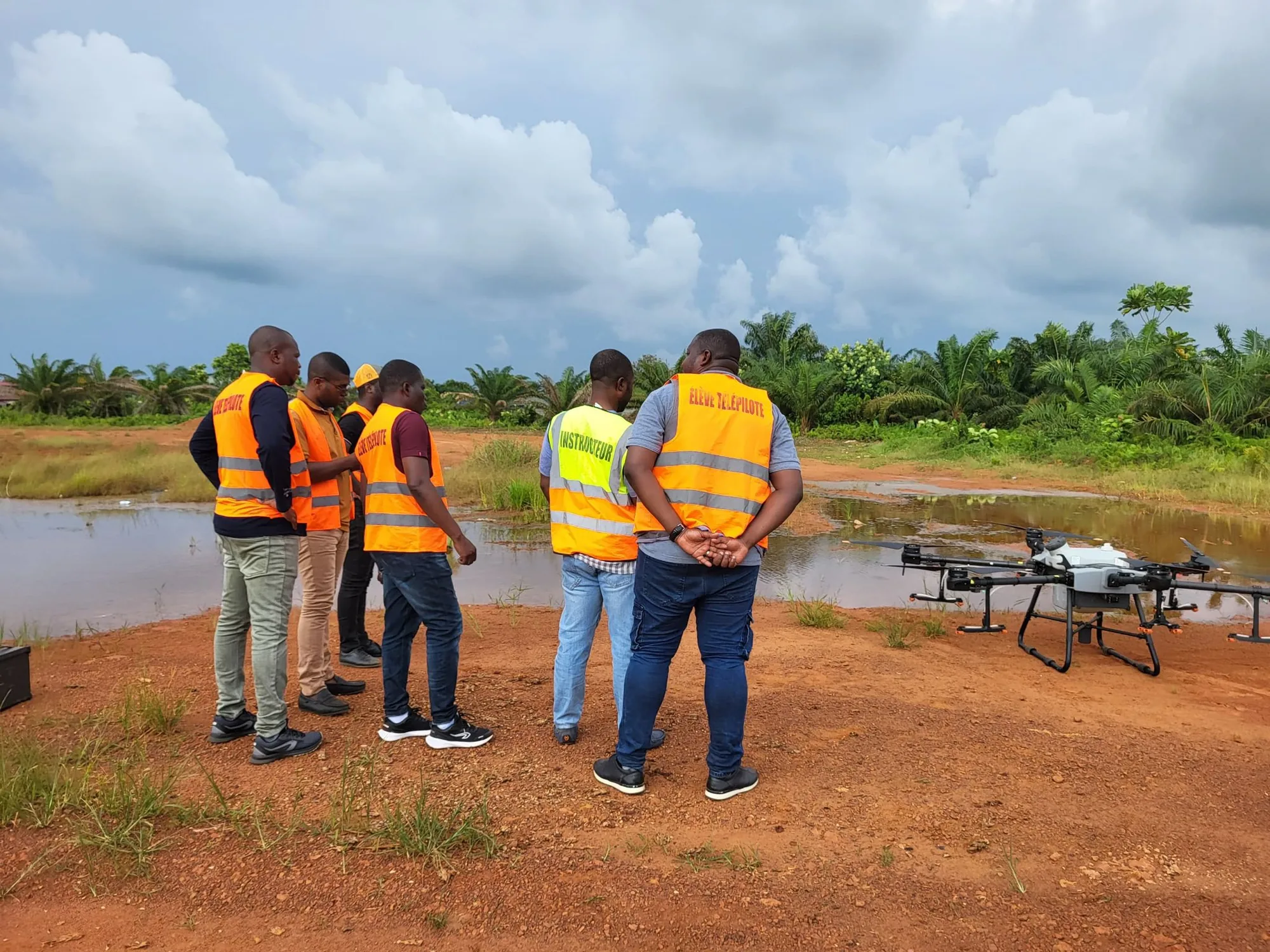 Stagiaires en gilet orange autour d'un drone d'épandage, au bord d'une parcelle inondée